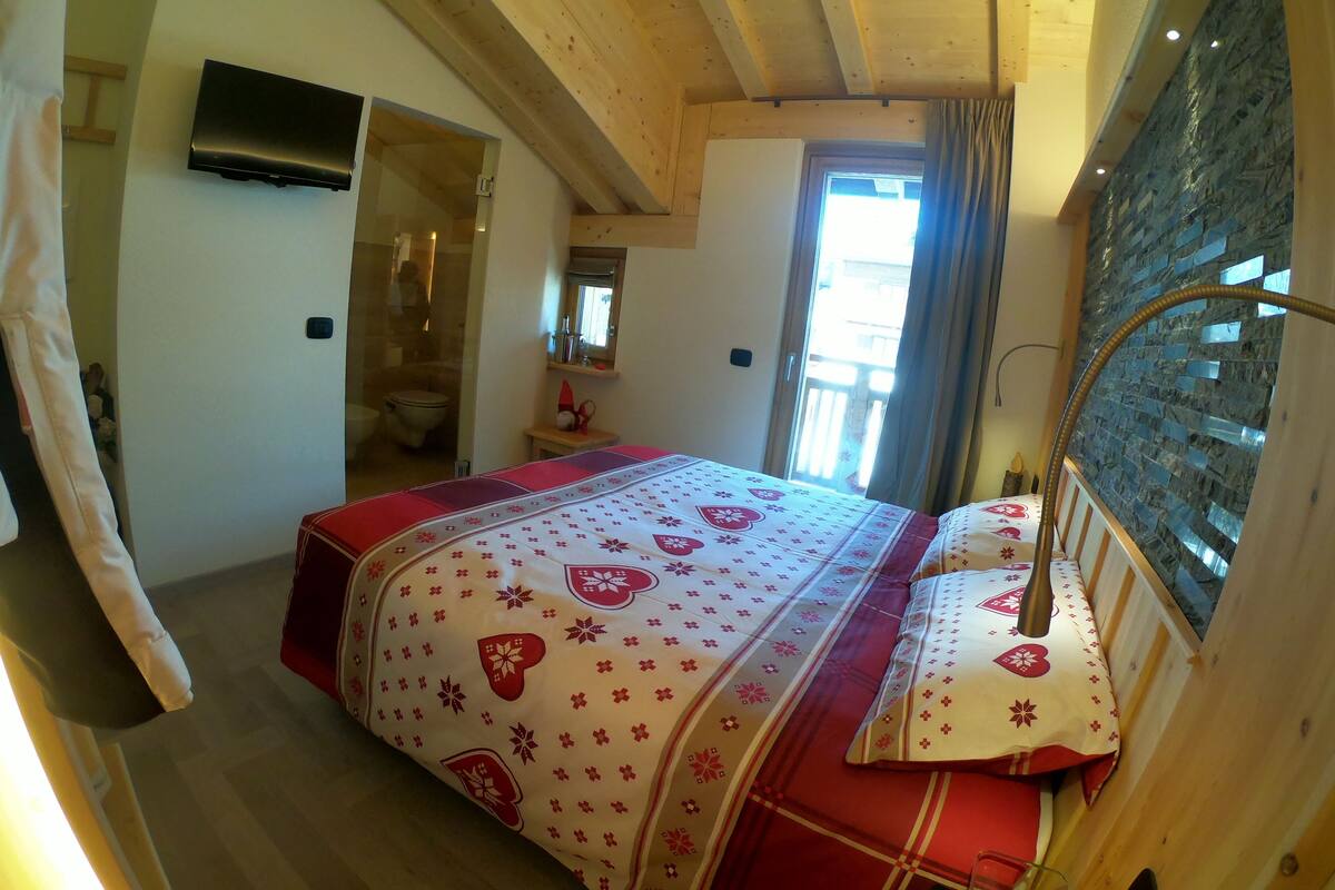 Ein Schlafzimmer mit Holzboden, einem Bett mit floralem Überwurf, einem durchgegangenen Tür sichtbar liegenden Badezimmer und einem Fenster mit Vorhängen.