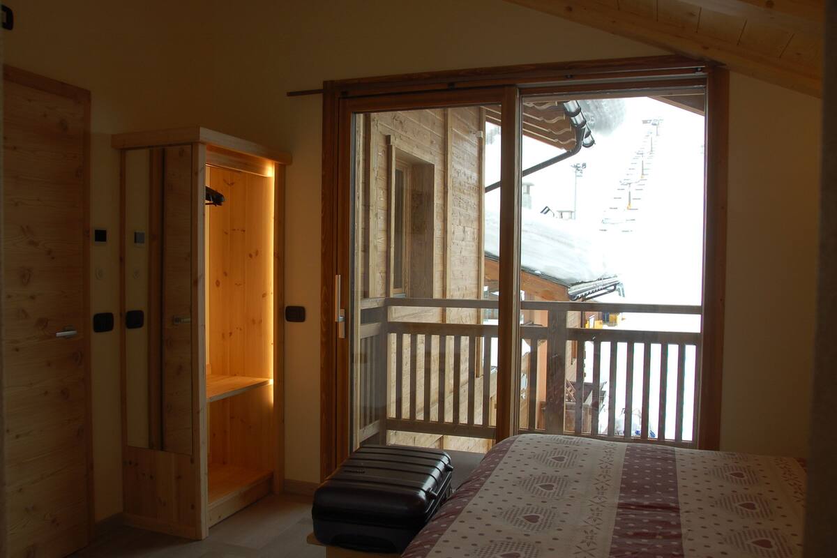 Ein Zimmer mit einem Bett, einem Koffer, einem Holzschrank und einer Glastür, die auf einen Balkon mit Blick auf eine verschneite Landschaft führt.