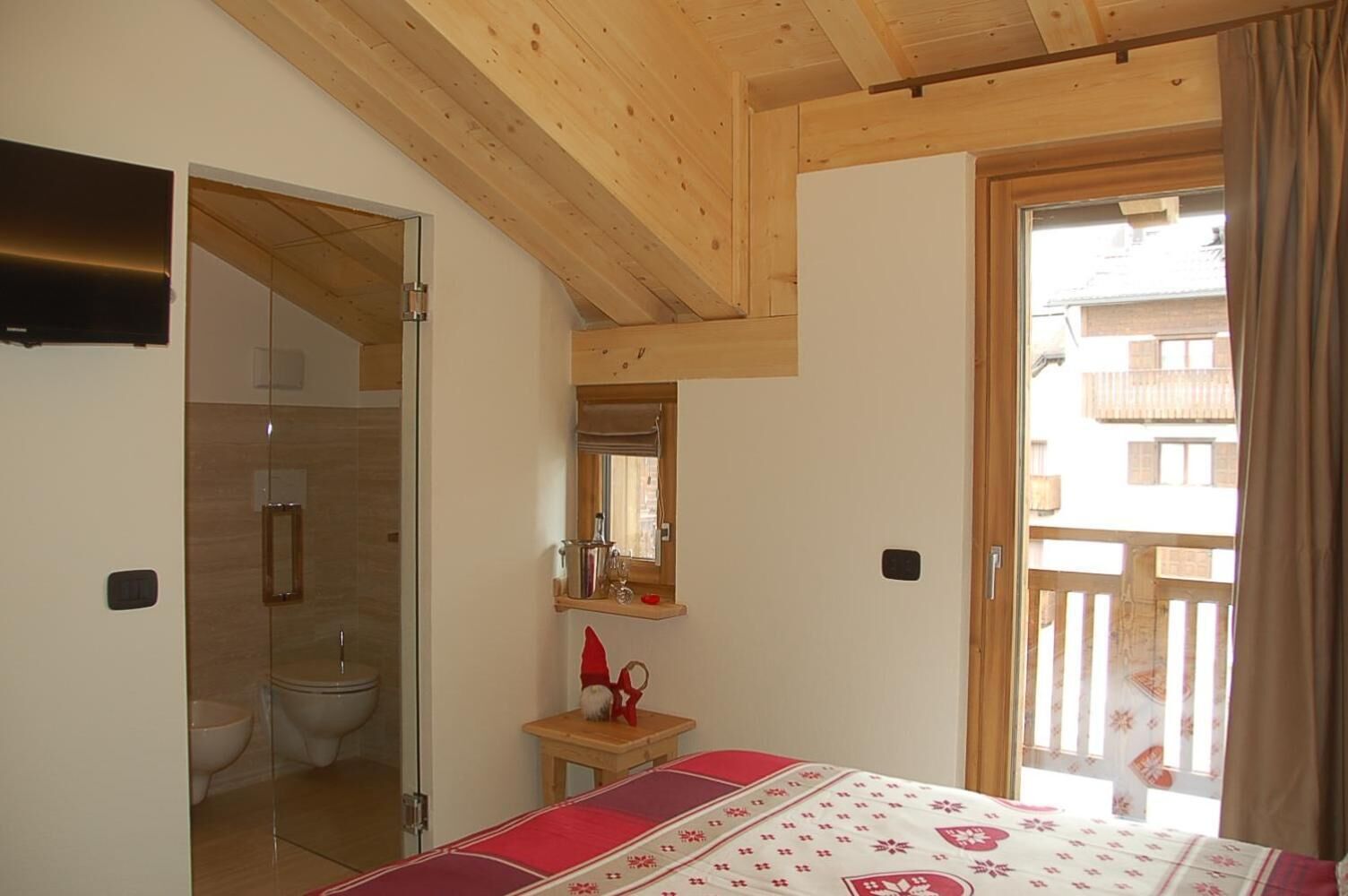 A bedroom with a wooden ceiling, a bed with a patterned bedspread, a small table with a red object, a mirror above the table, and a bathroom with a toilet and shower visible.