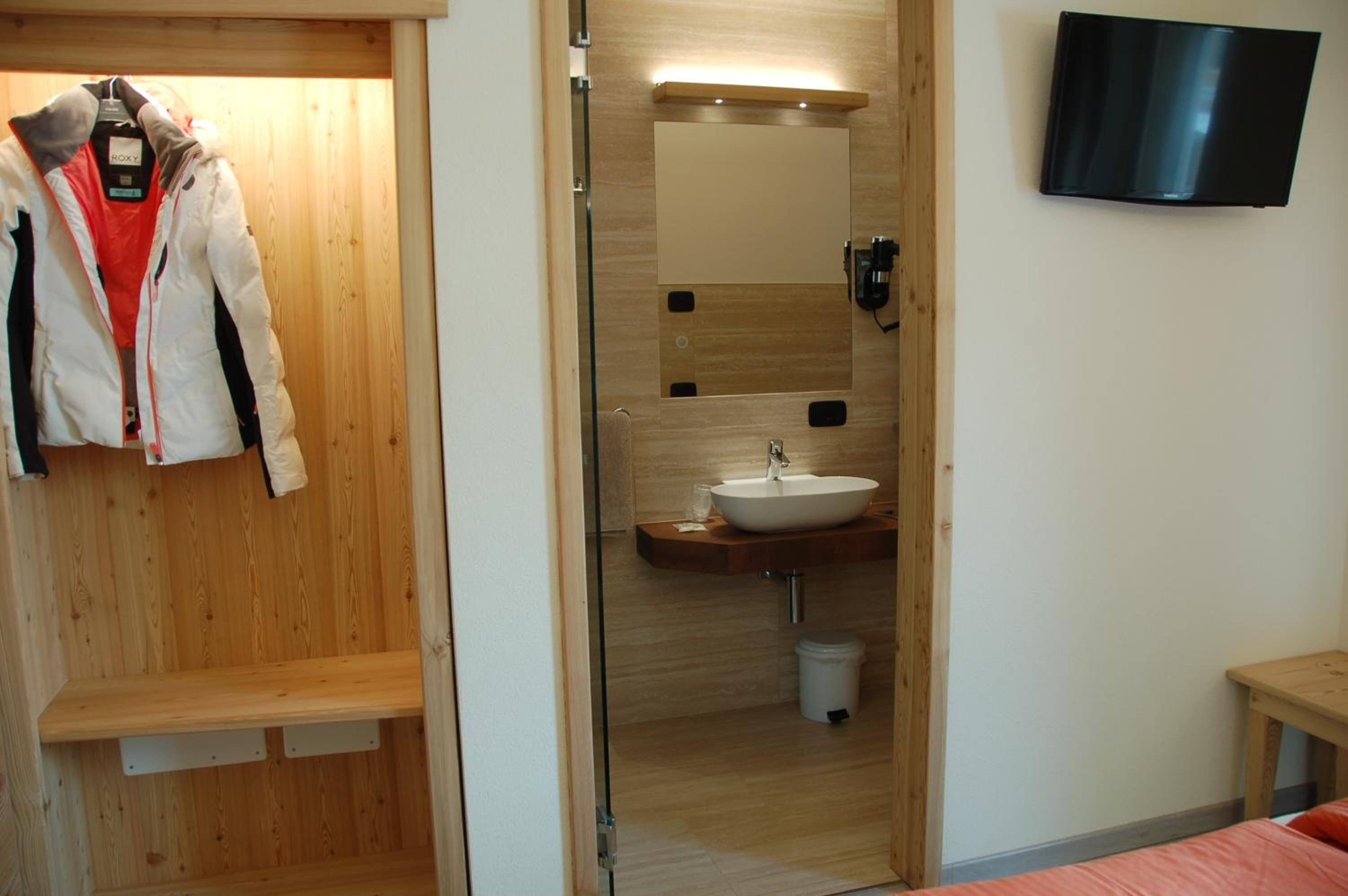 A hotel room with a wooden wardrobe, a mirror, a sink, and a television mounted on the wall.