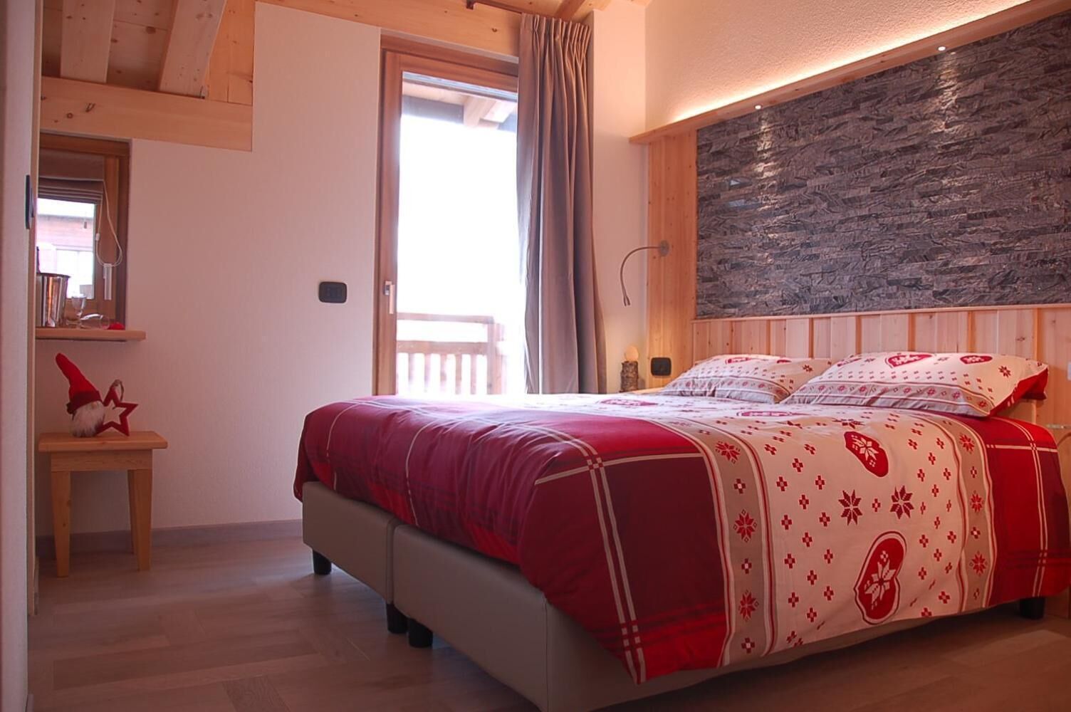 Quarto com cama, cabeceira de madeira, parede de pedra e uma mesinha com decoração de estrela.
