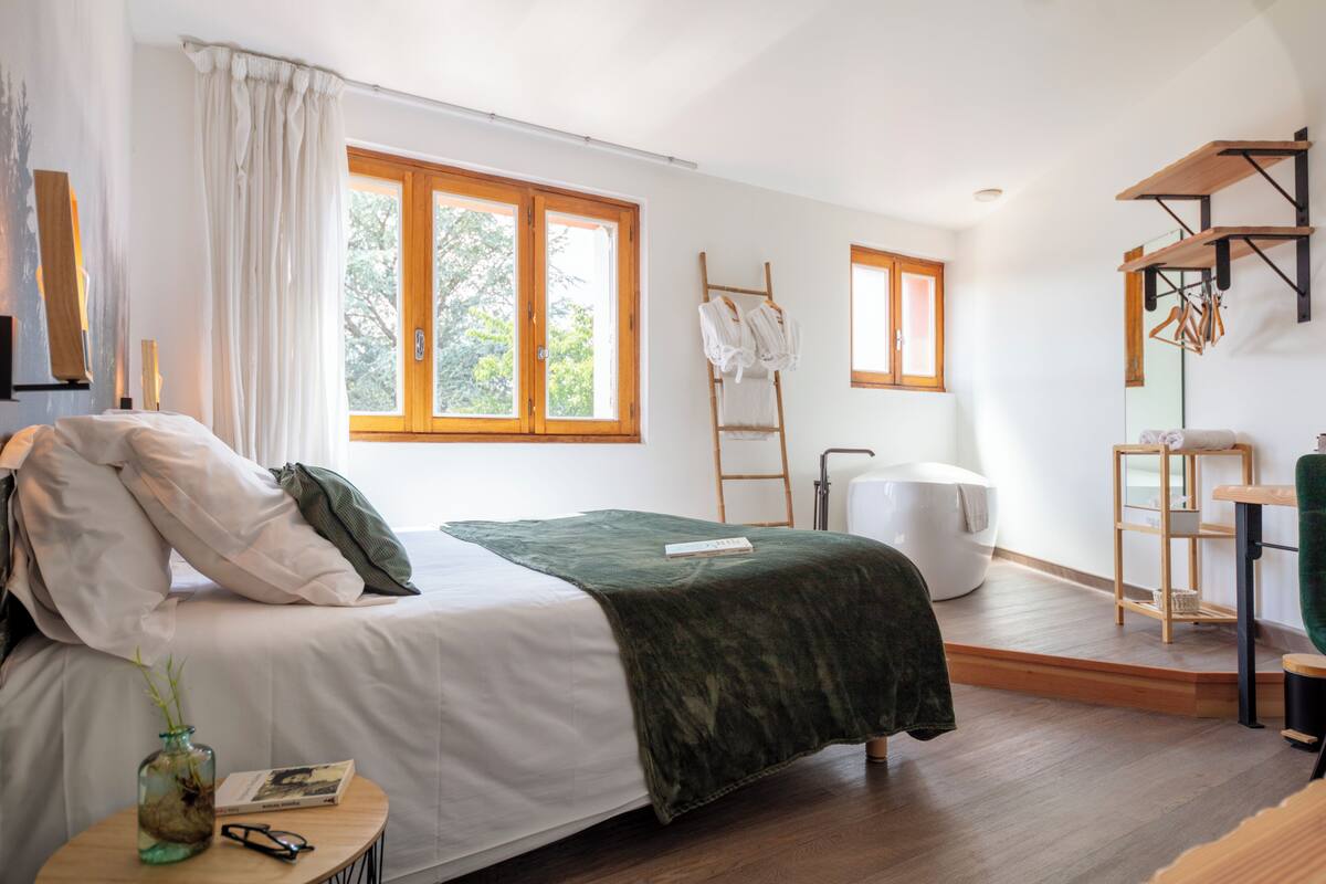 Un dormitorio moderno con una cama, un plaid verde, una mesita de noche con un libro y gafas, una escalera de madera y una silla blanca.