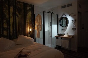 Un dormitorio con una cama grande, cabina de ducha de vidrio, espejo redondo y mesita de madera con lavamanos.