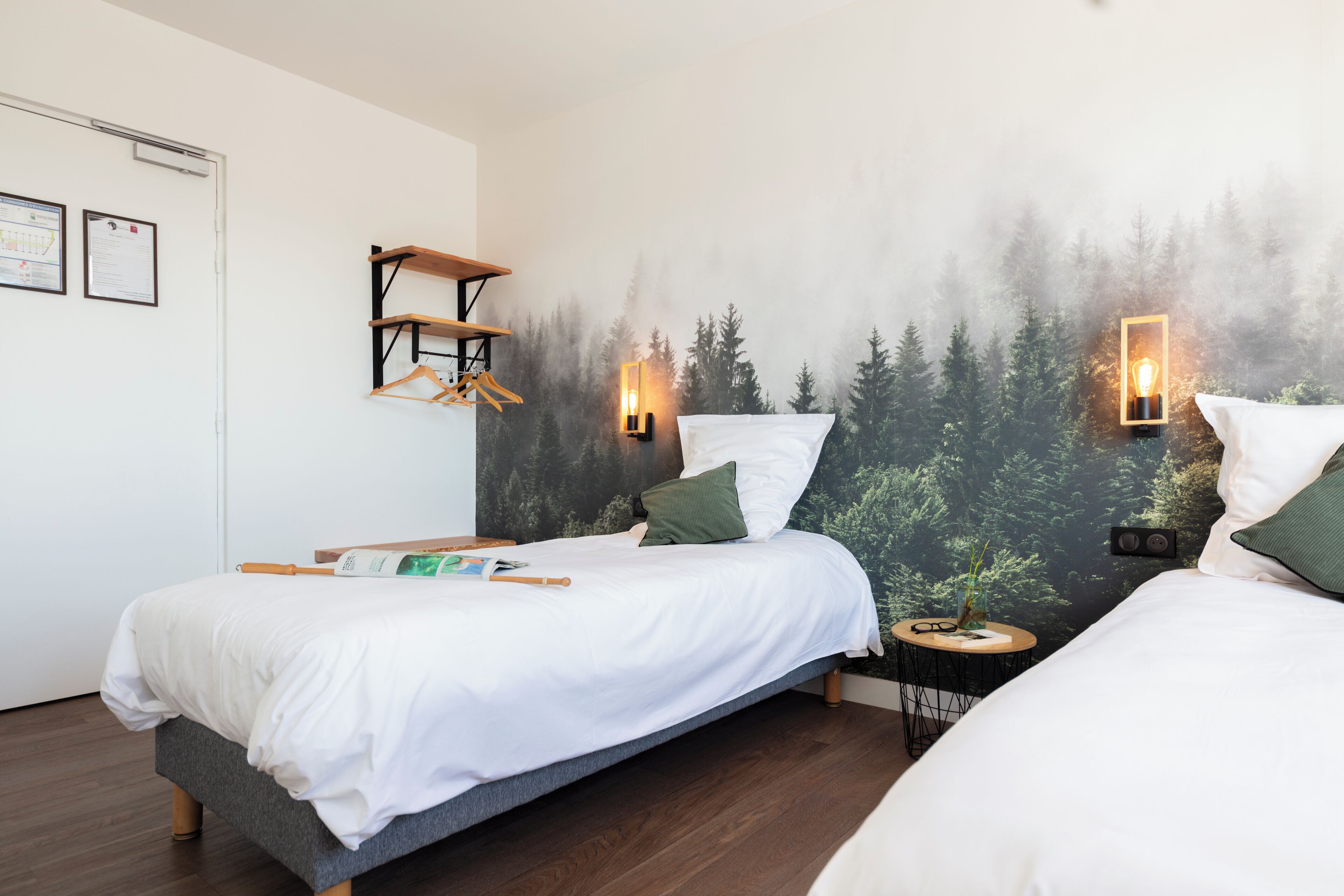 A bedroom with two beds, a wooden shelf, a small round table, and a wall mural of a forest.