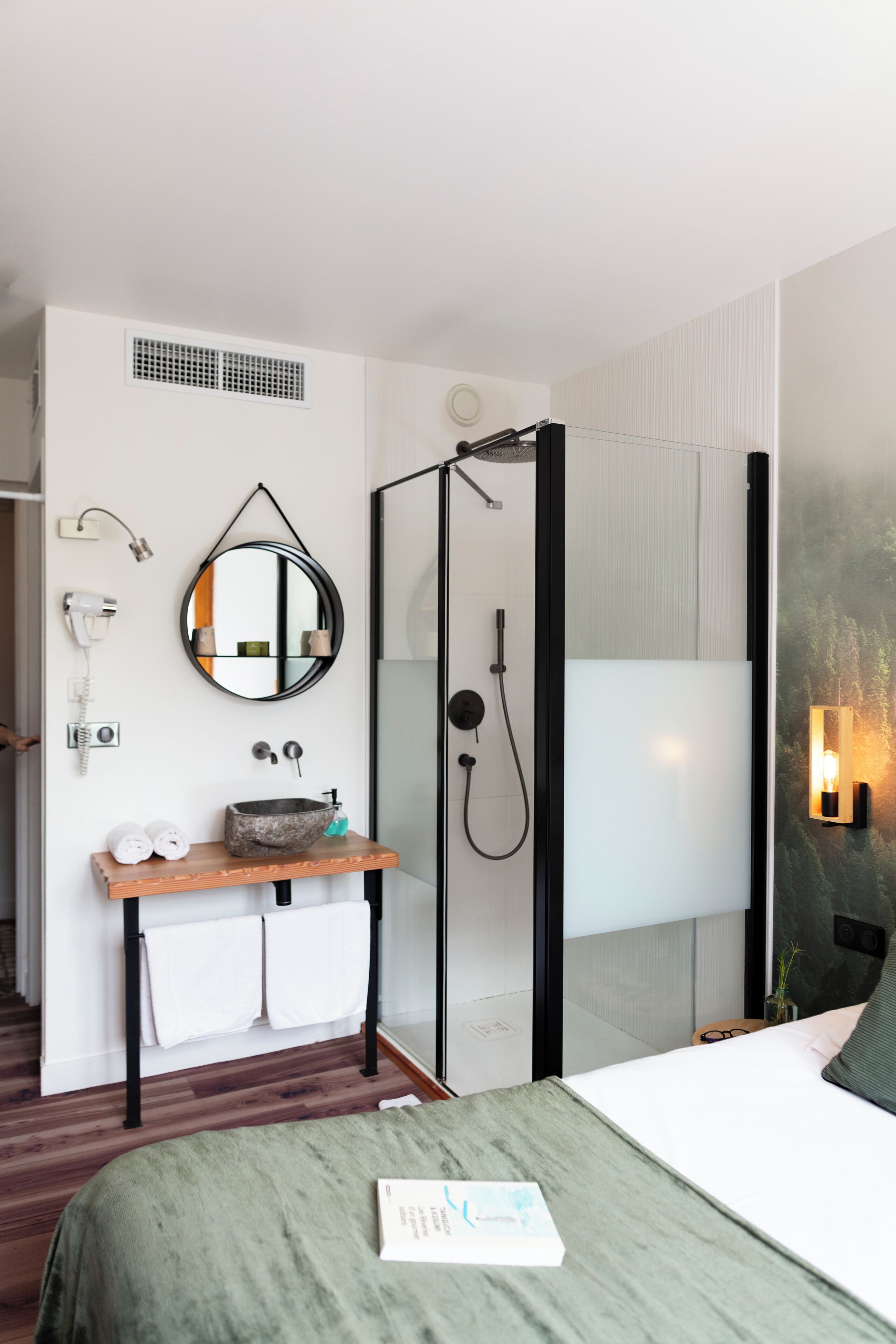 A modern hotel room with a wooden vanity, a round mirror, a glass shower enclosure, and a bed with a green bedspread.