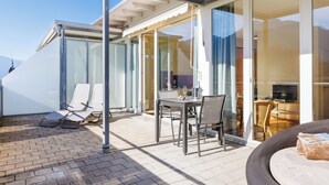 Une terrasse extérieure ensoleillée, avec une table et des chaises, un espace salon abrité et une porte vitrée donnant sur un espace intérieur.