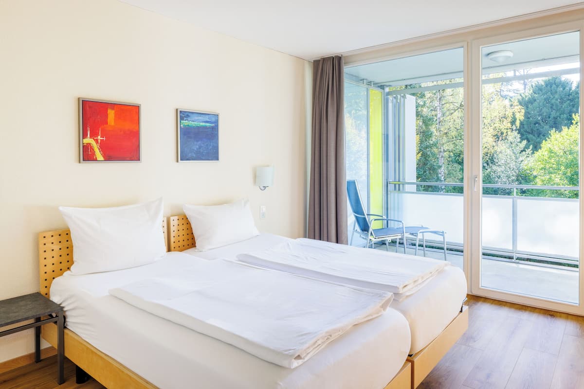A hotel room with a large bed, two paintings on the wall, a balcony with a chair, and a view of trees.