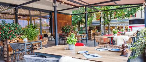 An outdoor cafe with wooden tables and wicker chairs, surrounded by potted plants and a covered seating area.