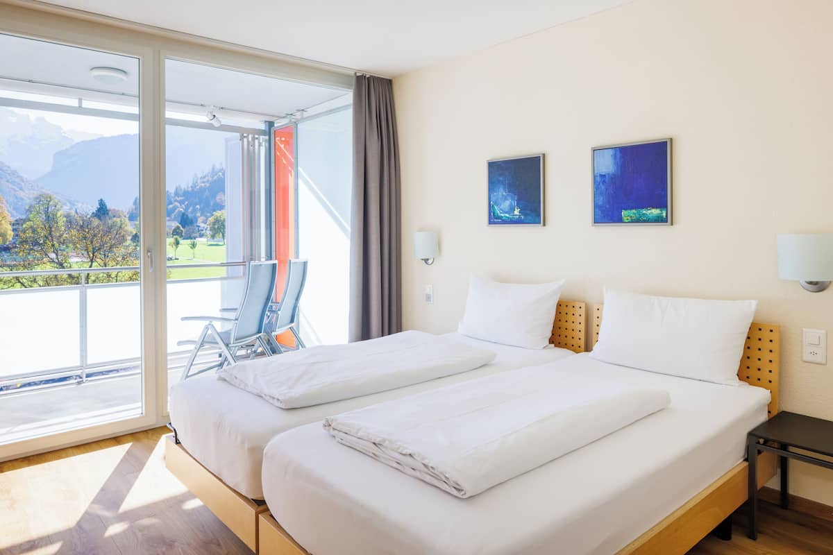 A hotel room with two beds, a balcony with a view of mountains, and two wall-mounted paintings.