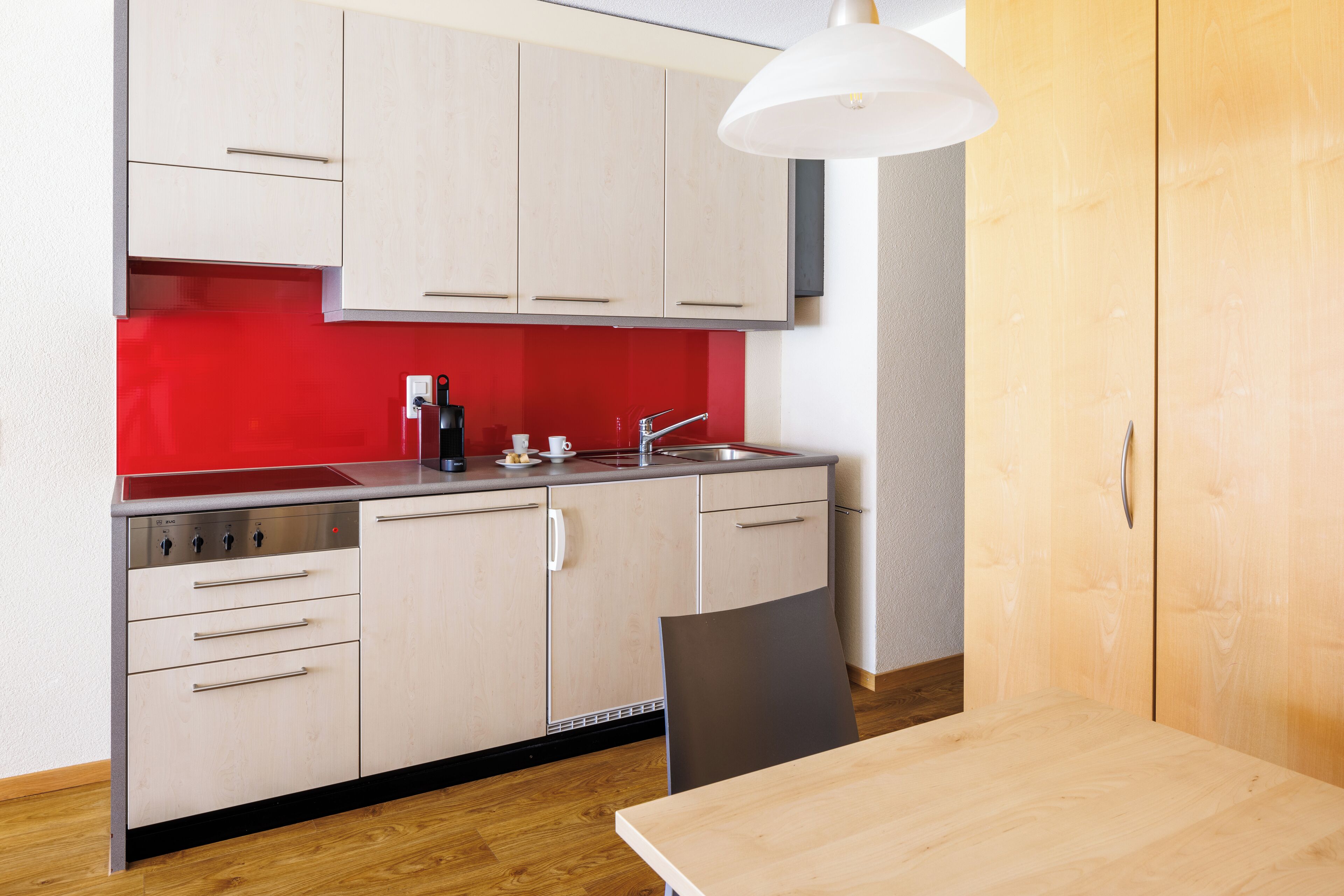 Cocina compacta con armarios de colores claros, salpicadero rojo y mesa de comedor de madera.
