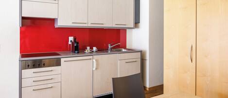 Cocina compacta con armarios de colores claros, salpicadero rojo y mesa de comedor de madera.