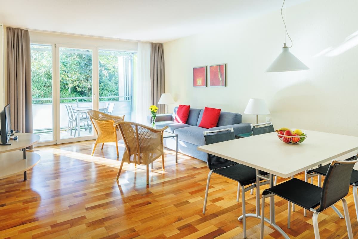 A modern living room with a sofa, dining table, and chairs. There is a balcony with a view of greenery.