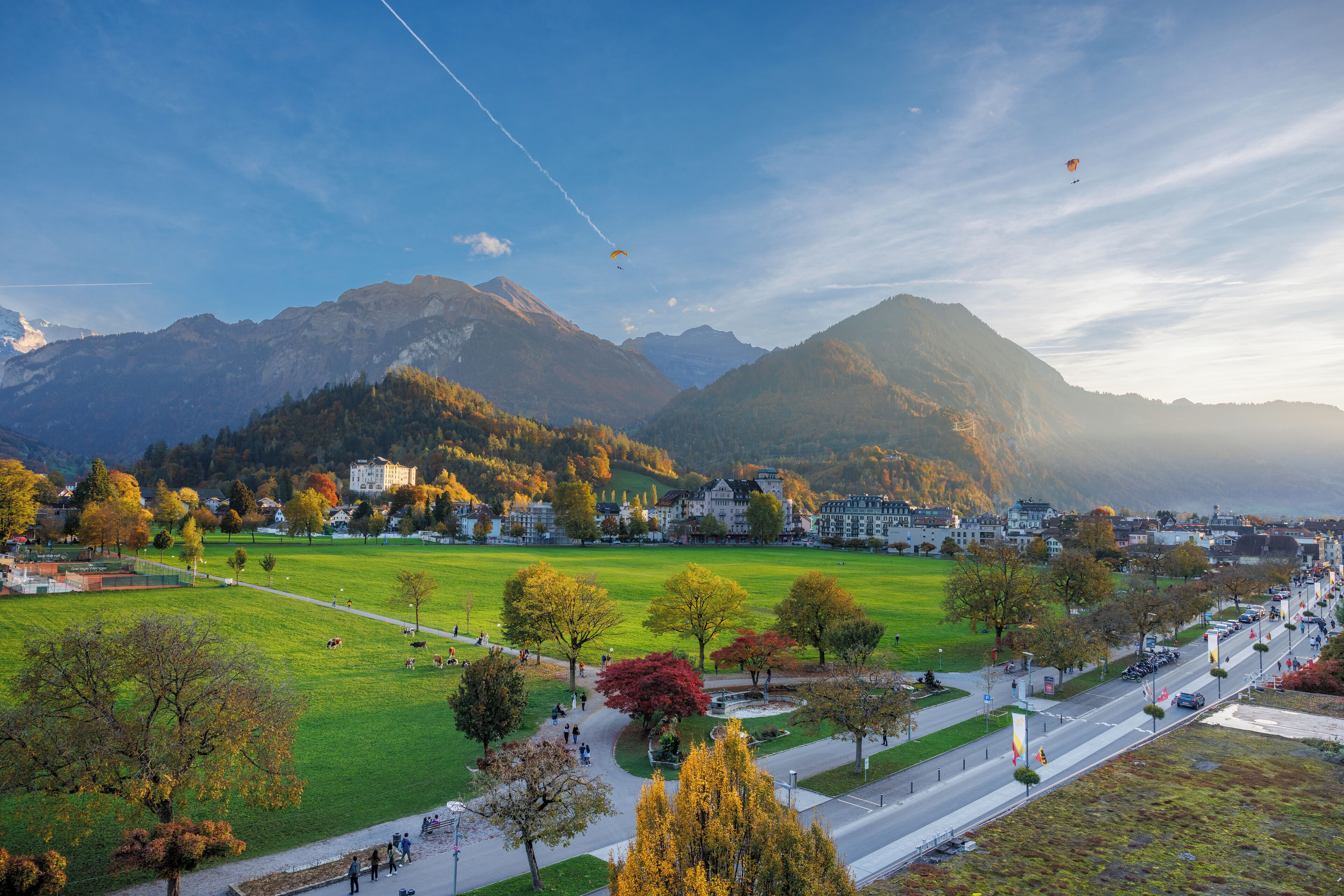 Une vue panoramique d’une ville avec un parc verdoyant, une route et des montagnes en arrière-plan.
