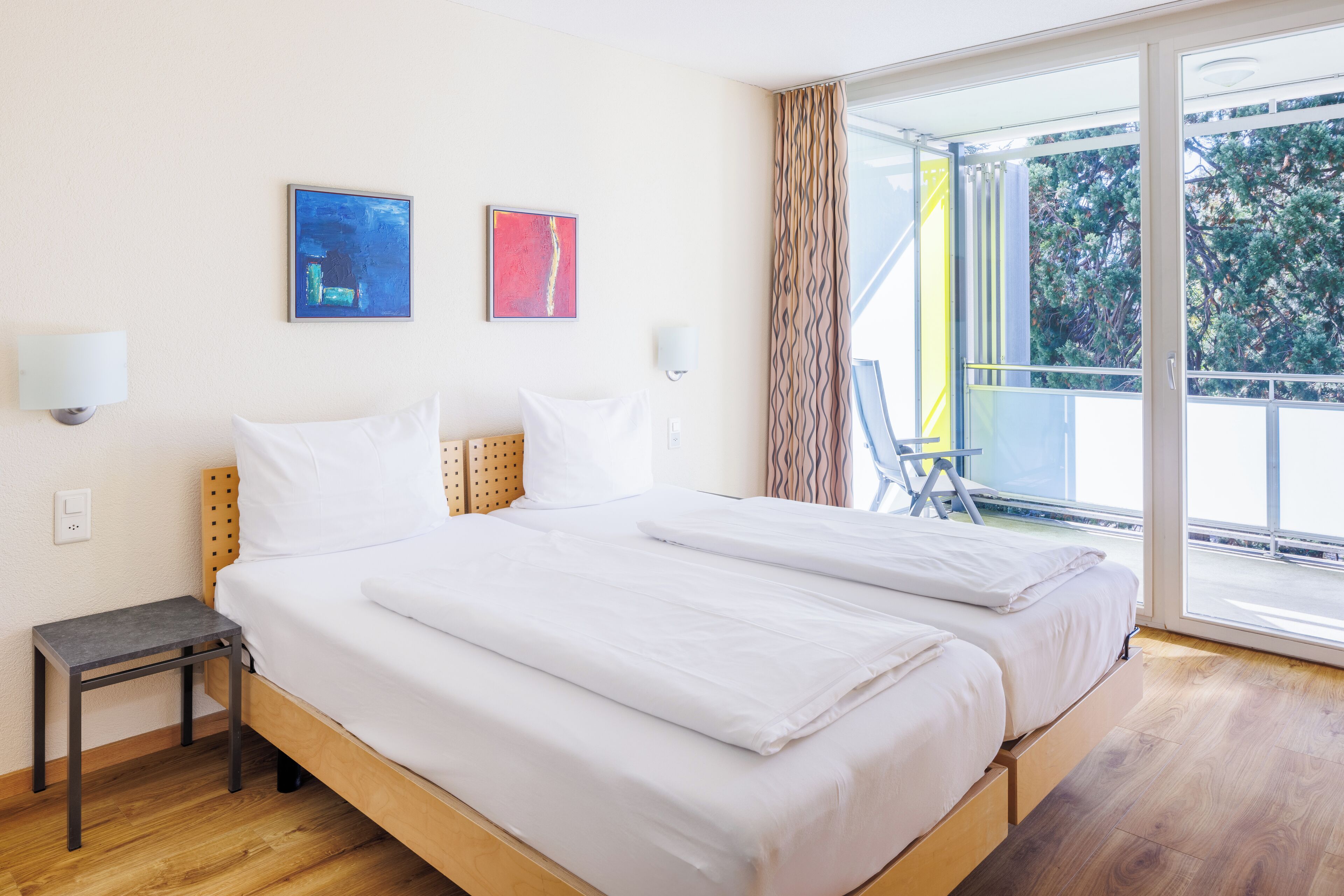 A hotel room with a large bed, two wall-mounted artworks, a small bedside table, and a view of the outdoors through a sliding glass door.