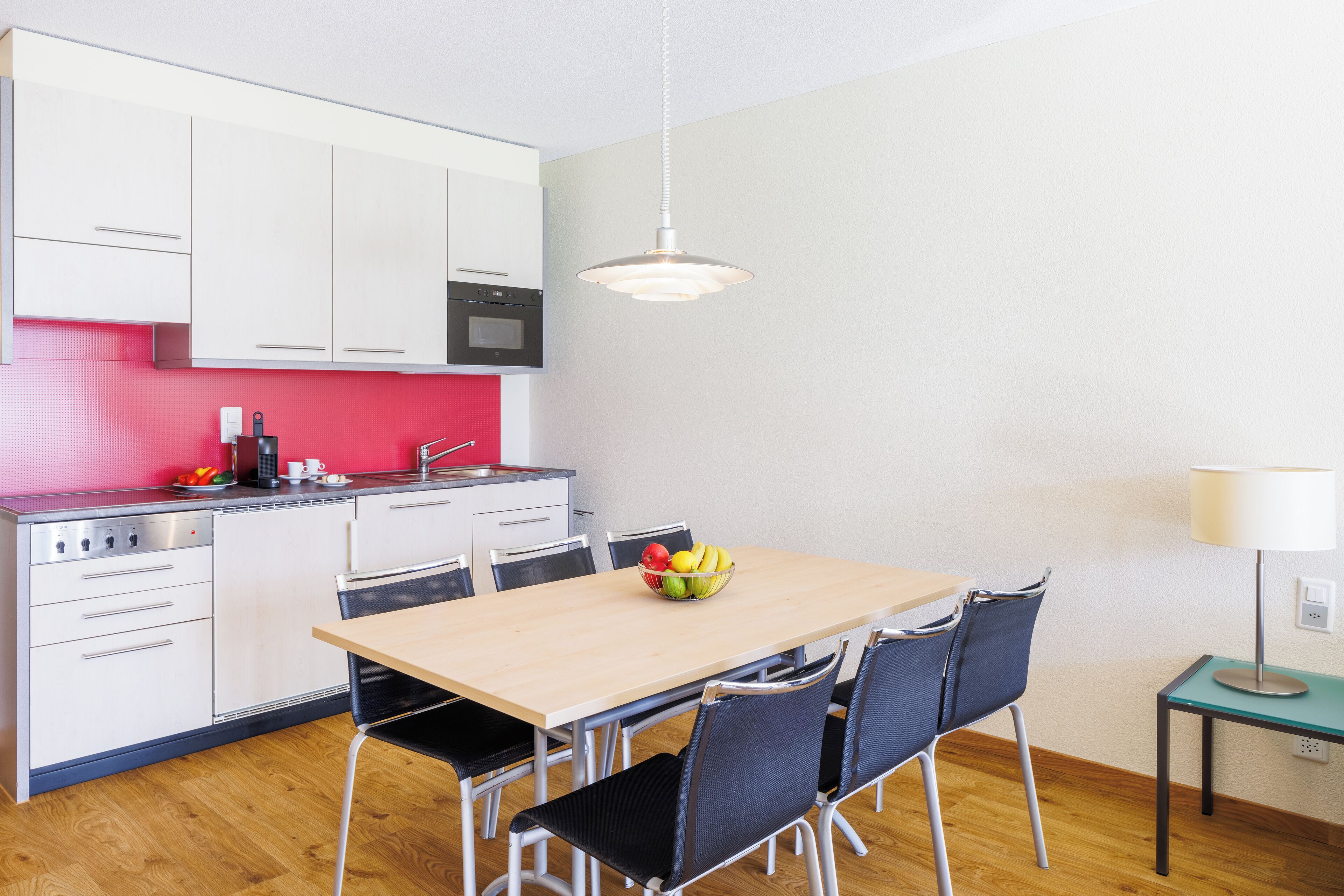 Une cuisine moderne avec des meubles blancs, un plan de travail rouge et une table à manger en bois, assortie de chaises.