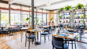 Un restaurant spacieux avec de hauts plafonds, de grandes fenêtres et des plantes d’intérieur. Les tables et les chaises sont disposées de manière ordonnée.