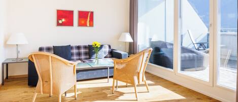 Ein modernes Wohnzimmer mit einer Couch, zwei Korbstühlen, einem Couchtisch und einem großen Fenster mit Blick auf Gartenmöbel.
