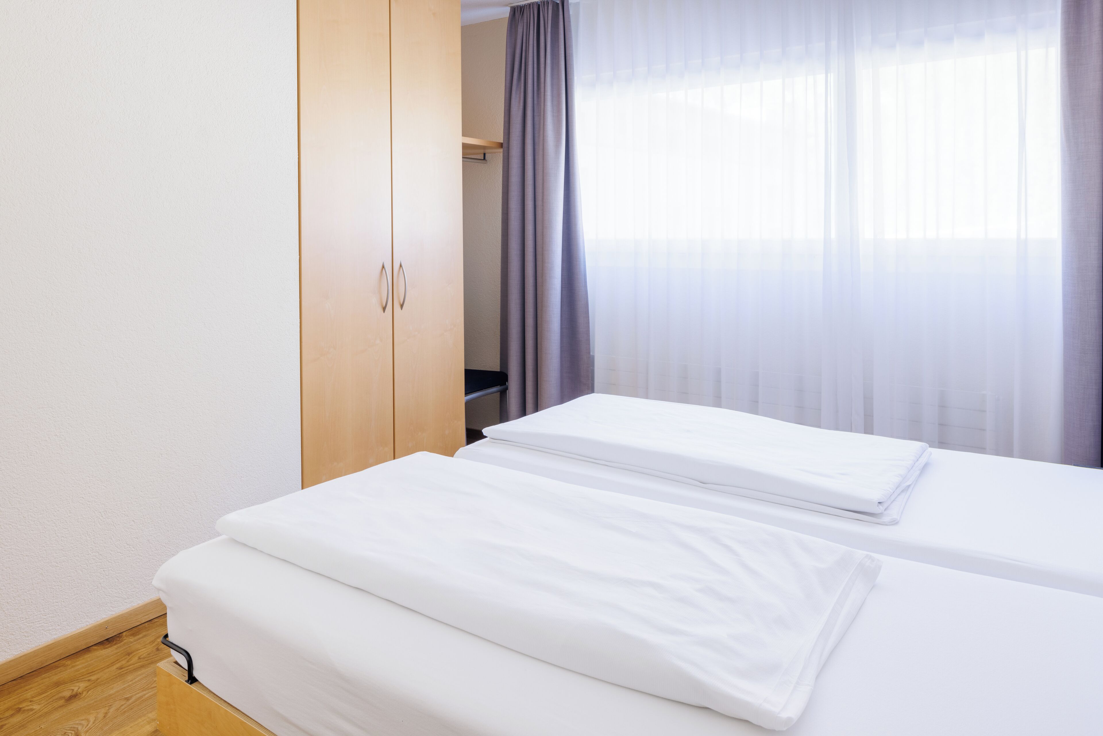 A hotel room with two single beds, a wooden wardrobe, and a window with sheer curtains.