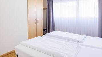 Una habitación de hotel con dos camas individuales, un armario de madera y una ventana con cortinas traslúcidas.