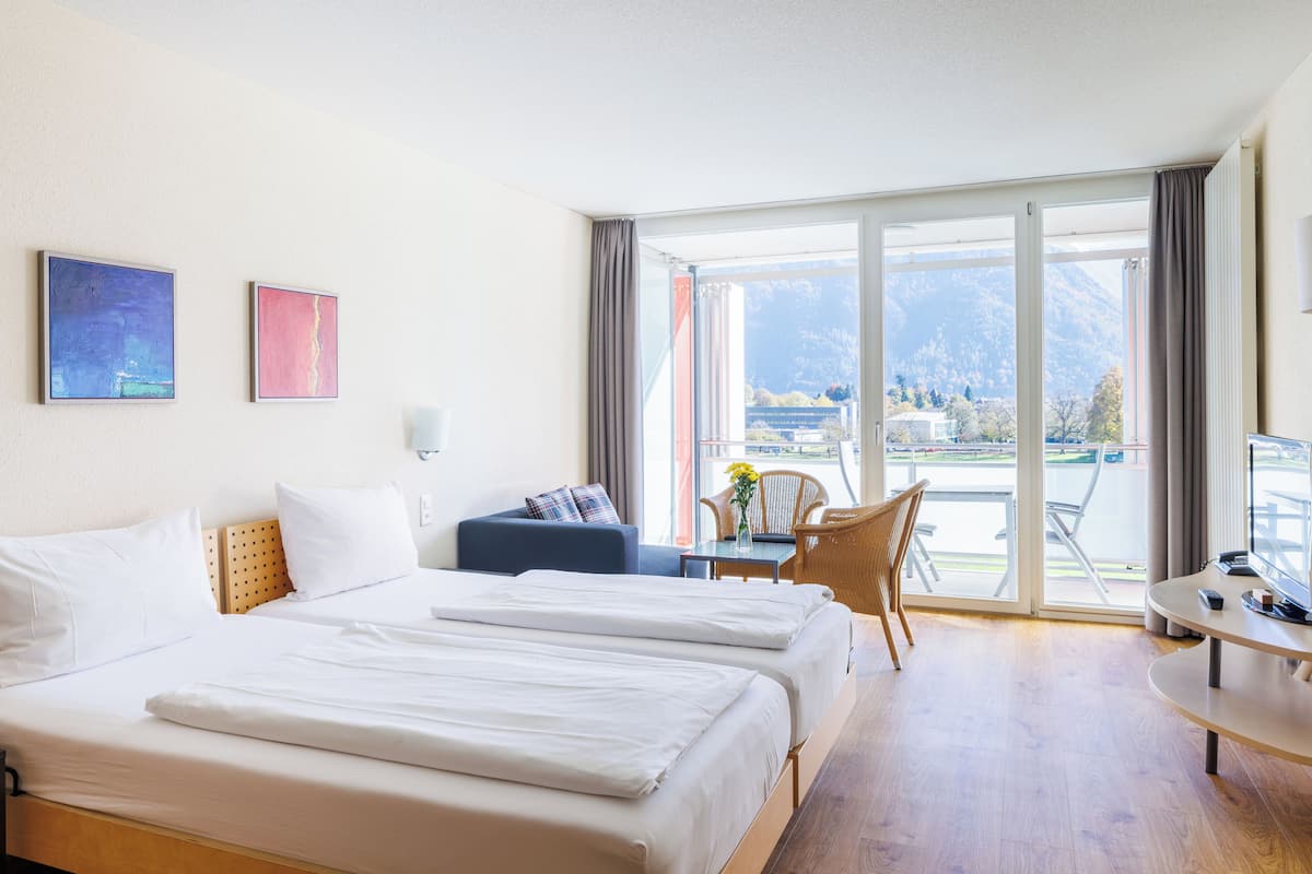 A hotel room with two beds, a sofa, a table, and a chair. There is a large window with a view of mountains and a balcony.