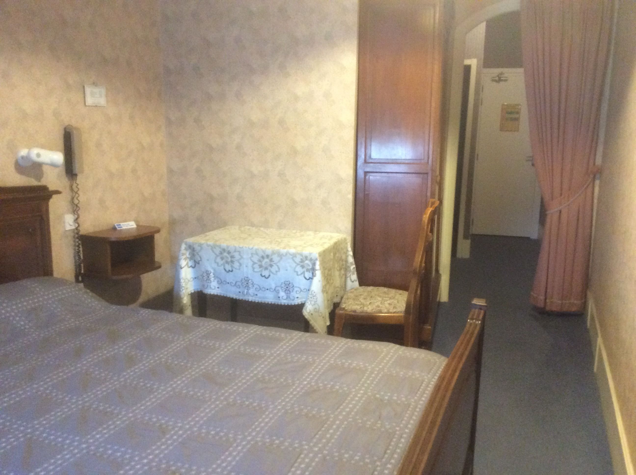 A hotel room with a bed, a small table with a tablecloth, a chair, a wooden wardrobe, and a door leading to another room.