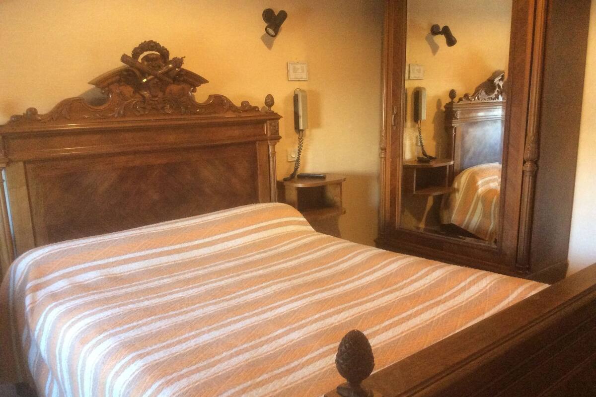 Un lit bien fait, recouvert d’une couvre-lit à rayures, avec une tête de lit en bois, une table de chevet sur laquelle se trouve un téléphone et un miroir qui reflète l’intérieur de la pièce.