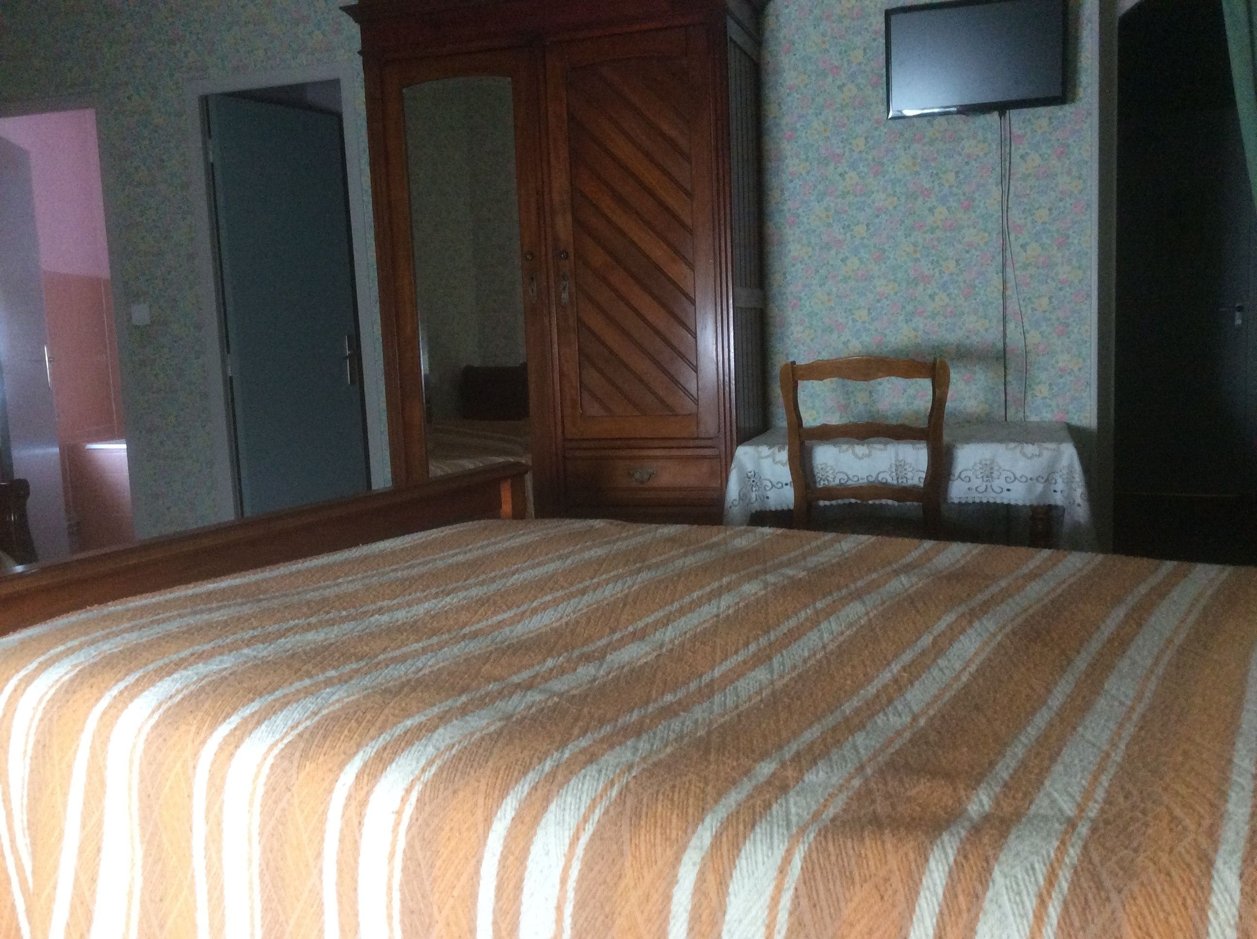 A bedroom with a large bed, a wooden wardrobe, a chair, a small table, and a television on the wall.