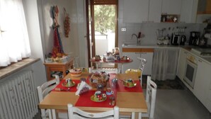 Un comedor con una mesa dispuesta para cuatro personas, rodeada de sillas blancas. Se observa un radiador a la izquierda, y una ventana con cortinas al fondo.