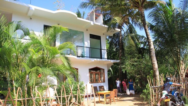 Une maison à deux étages avec un balcon, entourée de palmiers et bordée d’un chemin de terre.