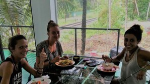 Trois personnes sont assises à une table dont le plateau est en verre, entourée de grandes fenêtres offrant une vue sur des palmiers et une végétation luxuriante.