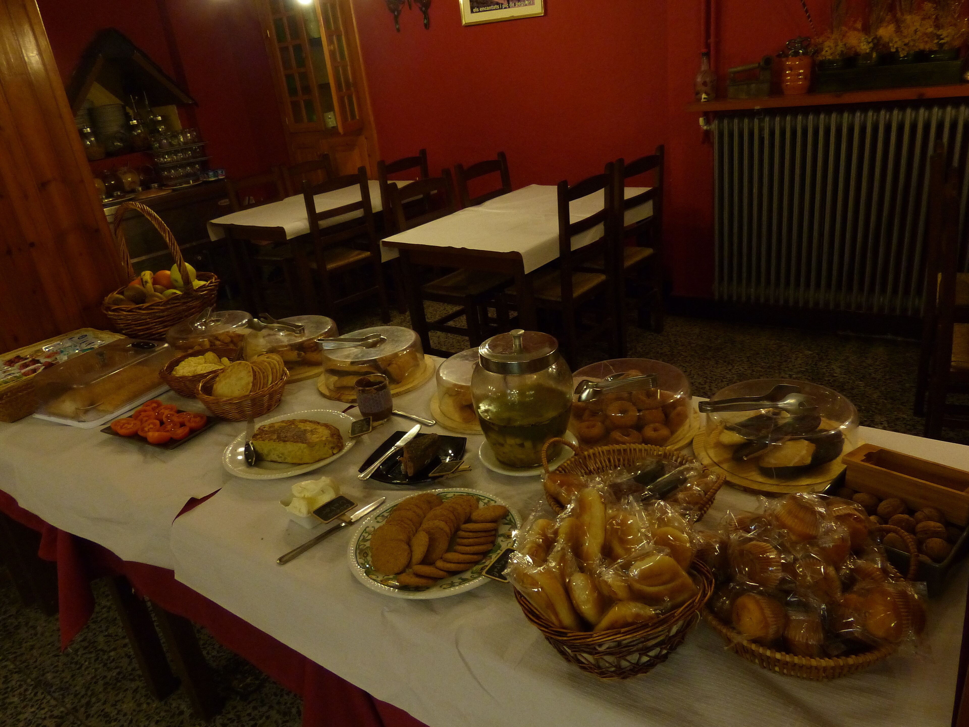 Mesa de jantar posta com diversos alimentos e utensílios, rodeada por cadeiras de madeira.