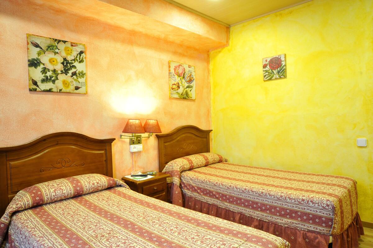 A hotel room with two beds, a nightstand, a lamp, and floral wall art.