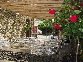 Un café al aire libre con mesas y sillas de metal, un muro de piedra y una pérgola de madera.