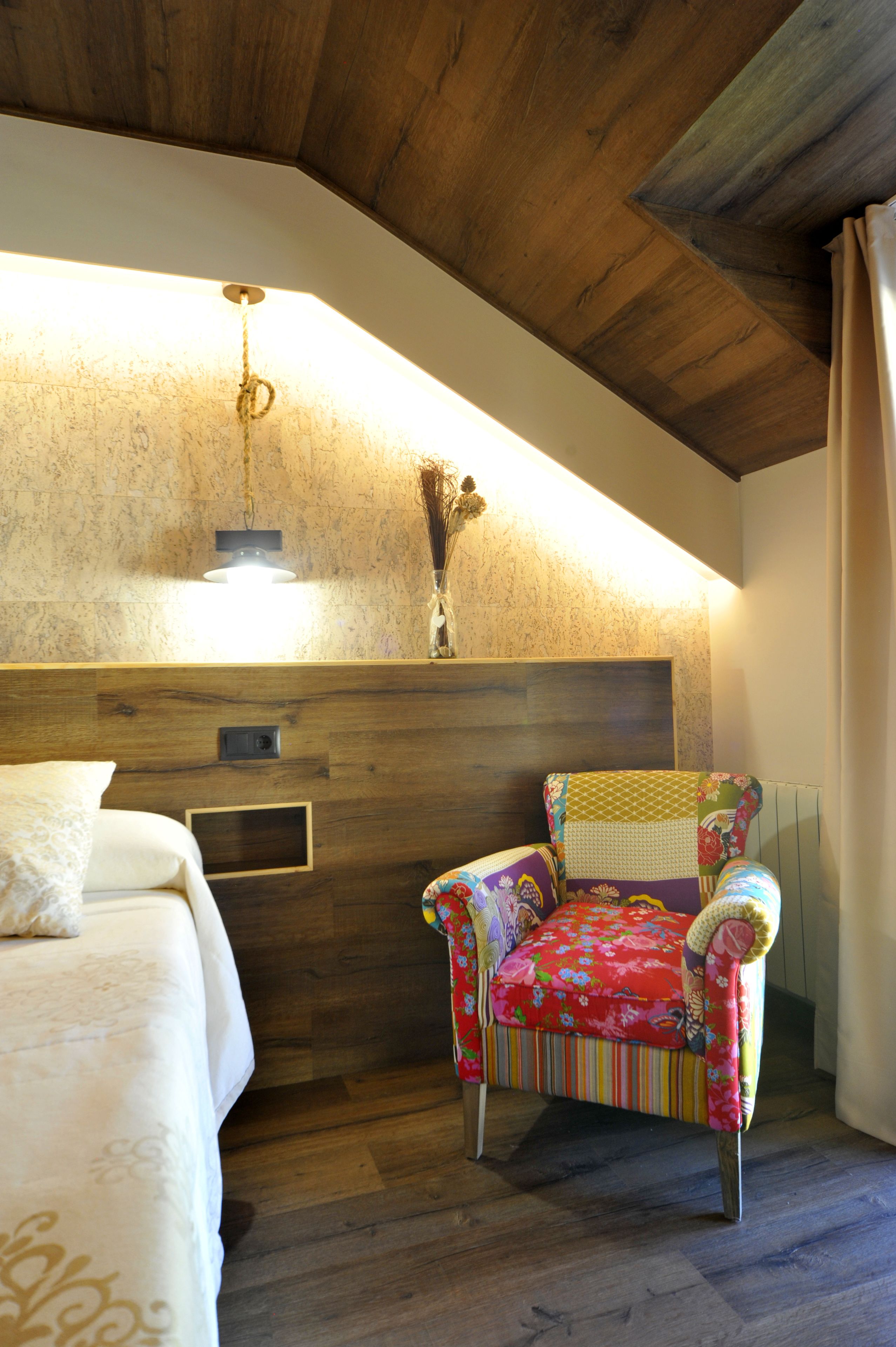 Une chambre avec un plafond en bois, un lit avec une literie blanche, une chaise colorée et un luminaire fixé au mur.