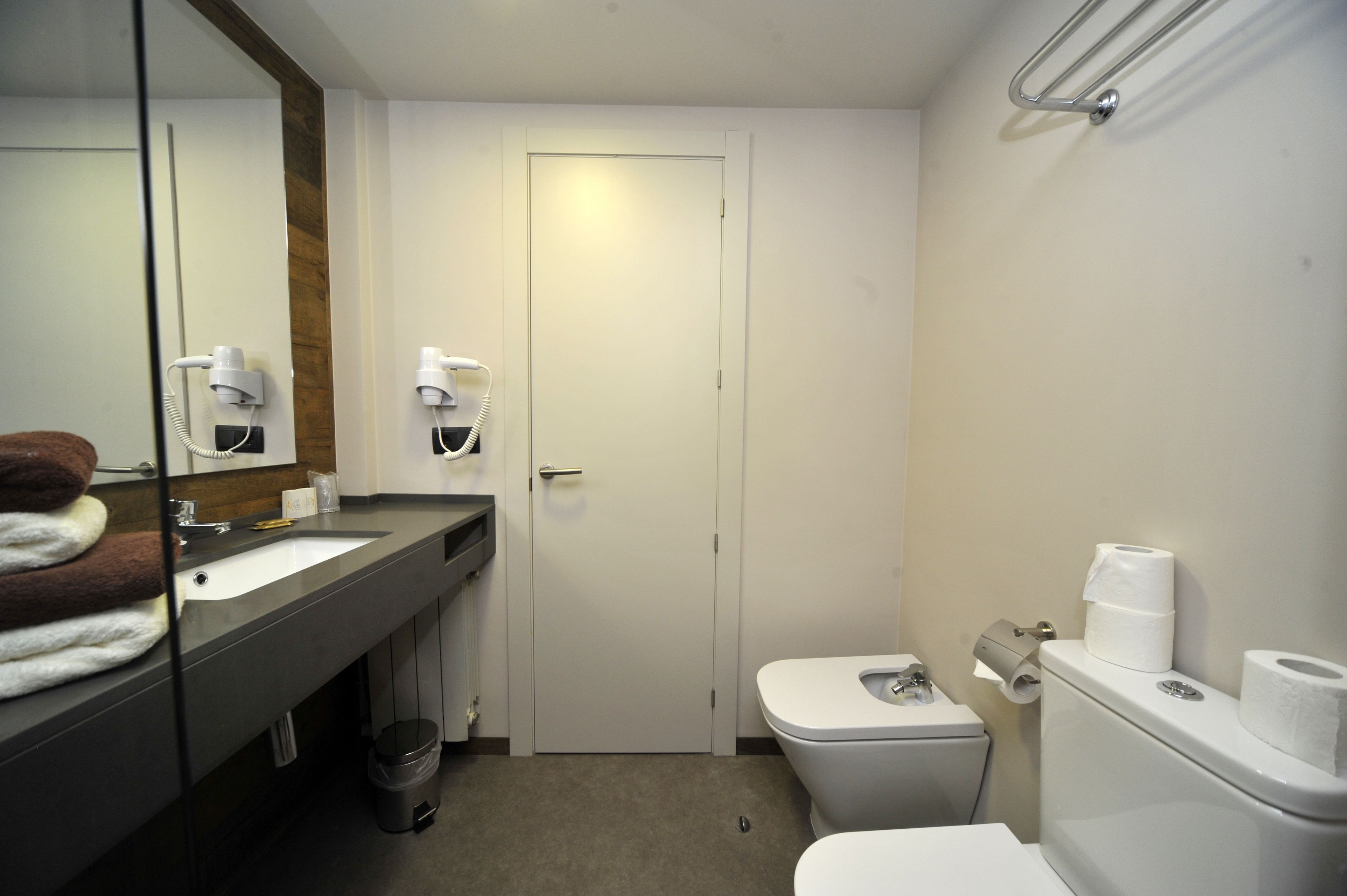 Un baño de hotel pequeño y funcional, con lavamanos, espejo, toallas apiladas, inodoro y puerta.