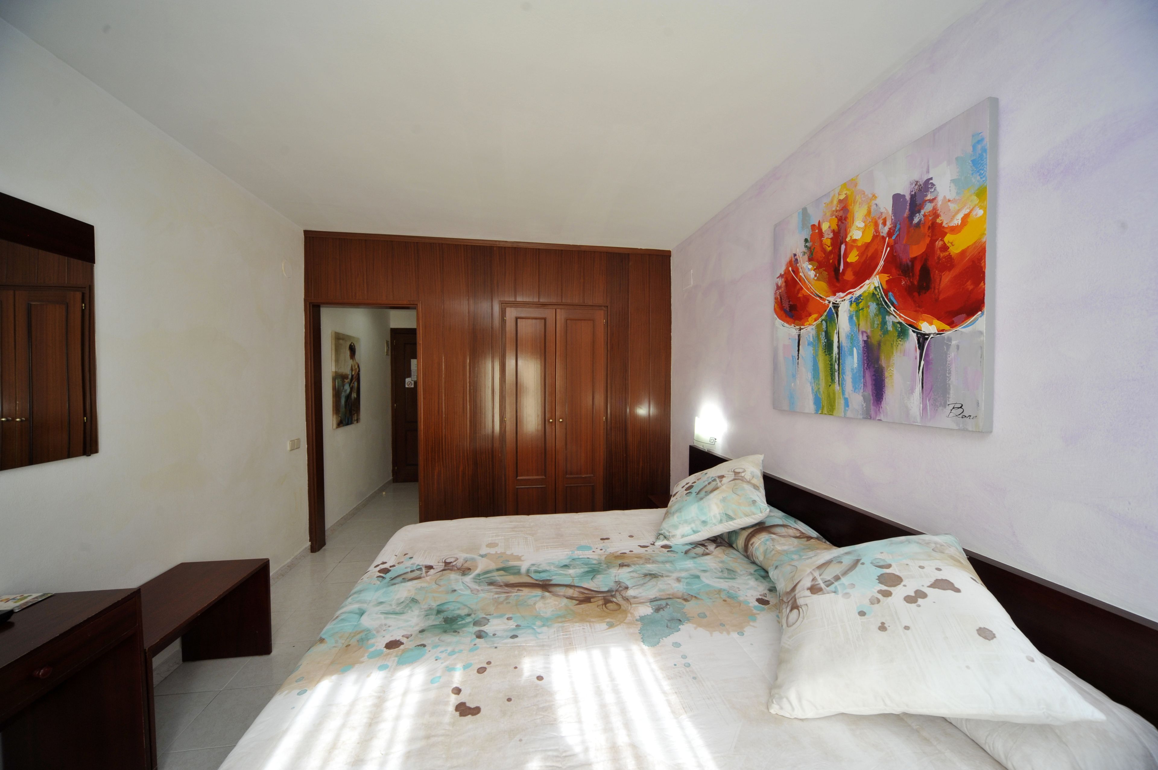 A bedroom with a wooden wardrobe, a bed with floral bedding, and a colorful abstract painting on the wall.