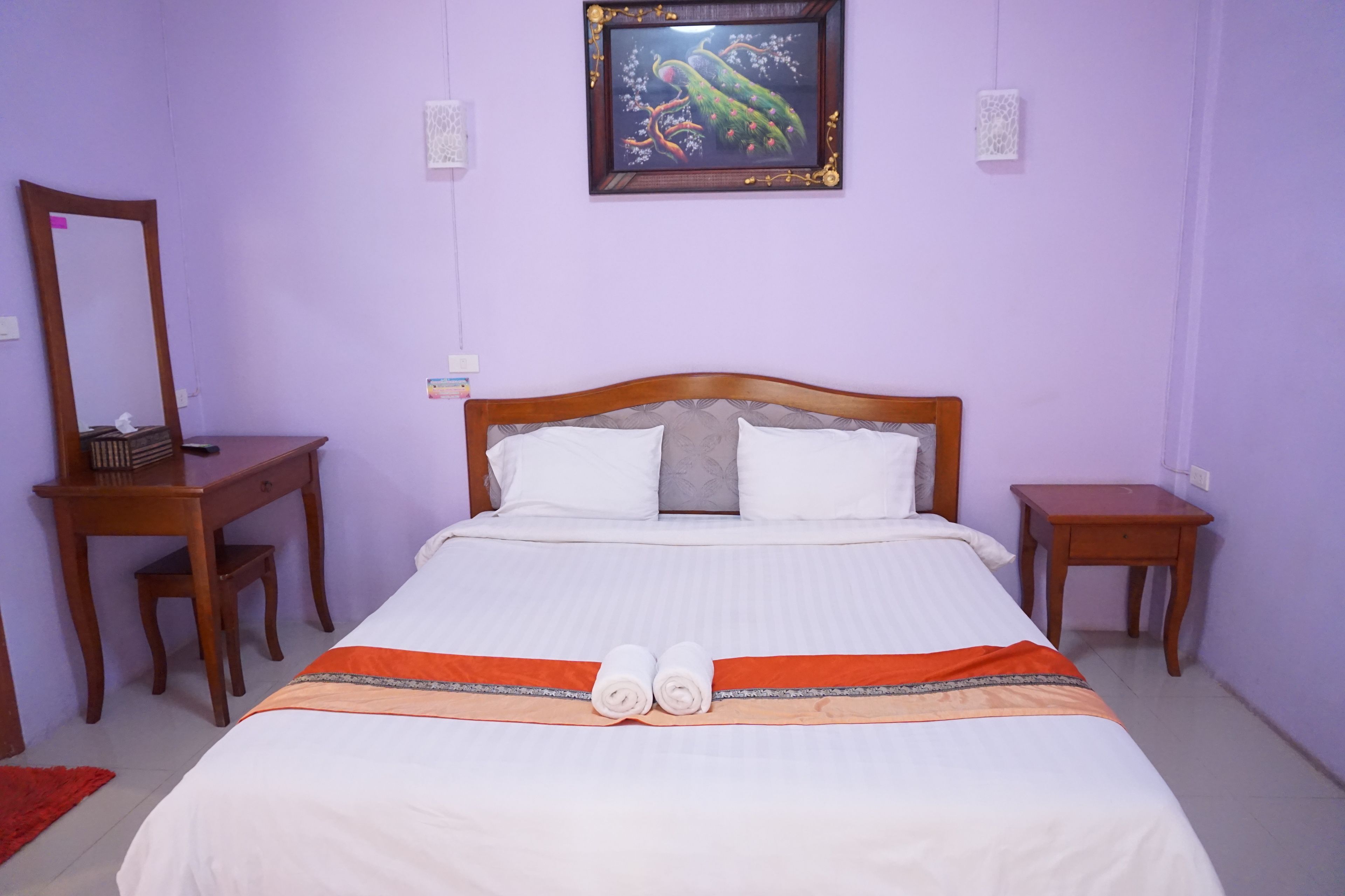 Una cama bien hecha con sábanas blancas y un detalle decorativo naranja, una mesita de noche de madera con una lámpara, un escritorio de madera con una silla y un cuadro en la pared.