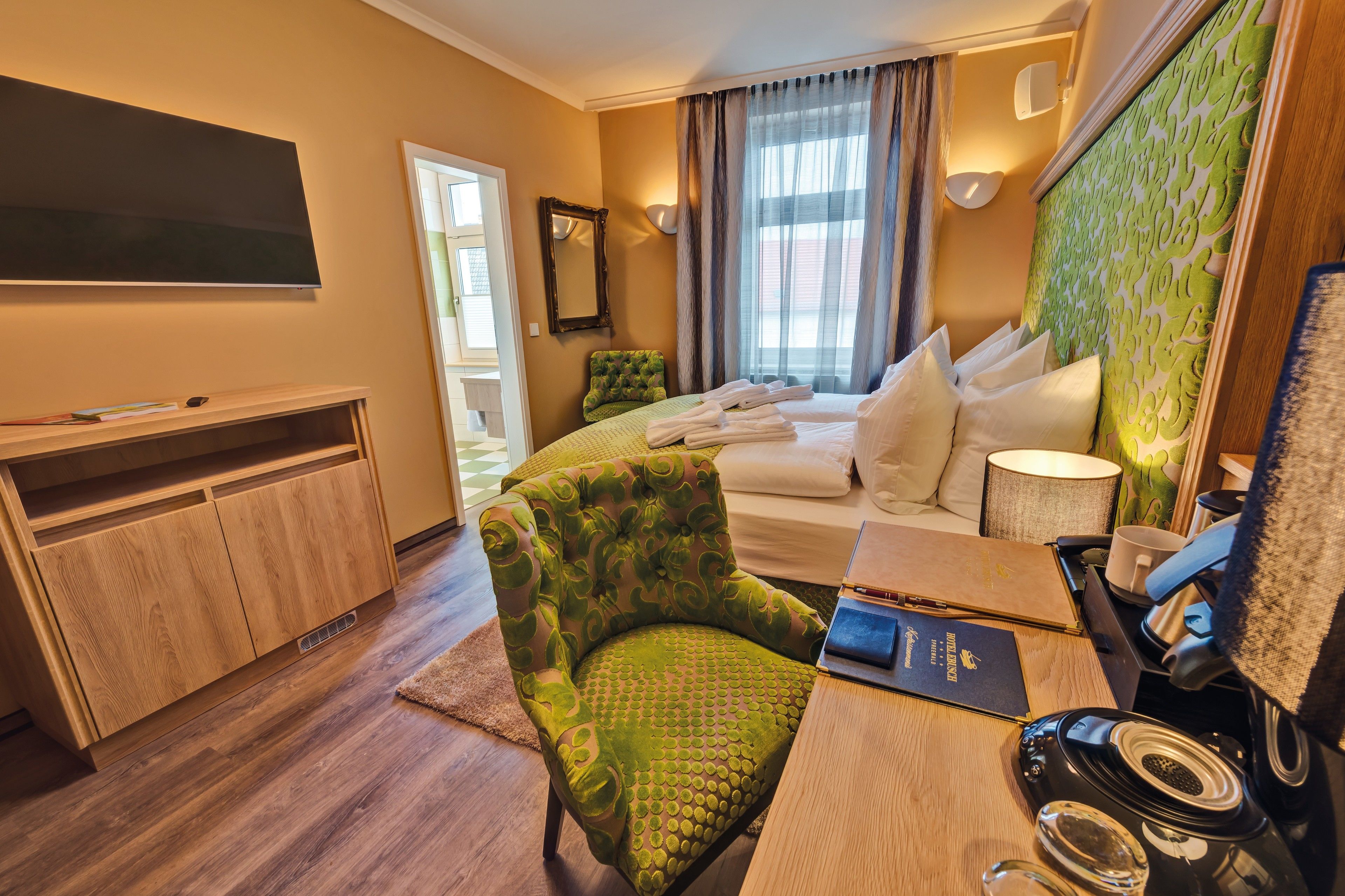 Habitación de hotel con cama, un sillón verde, un escritorio con una computadora portátil, un televisor de pantalla plana y un gran ventanal con cortinas.