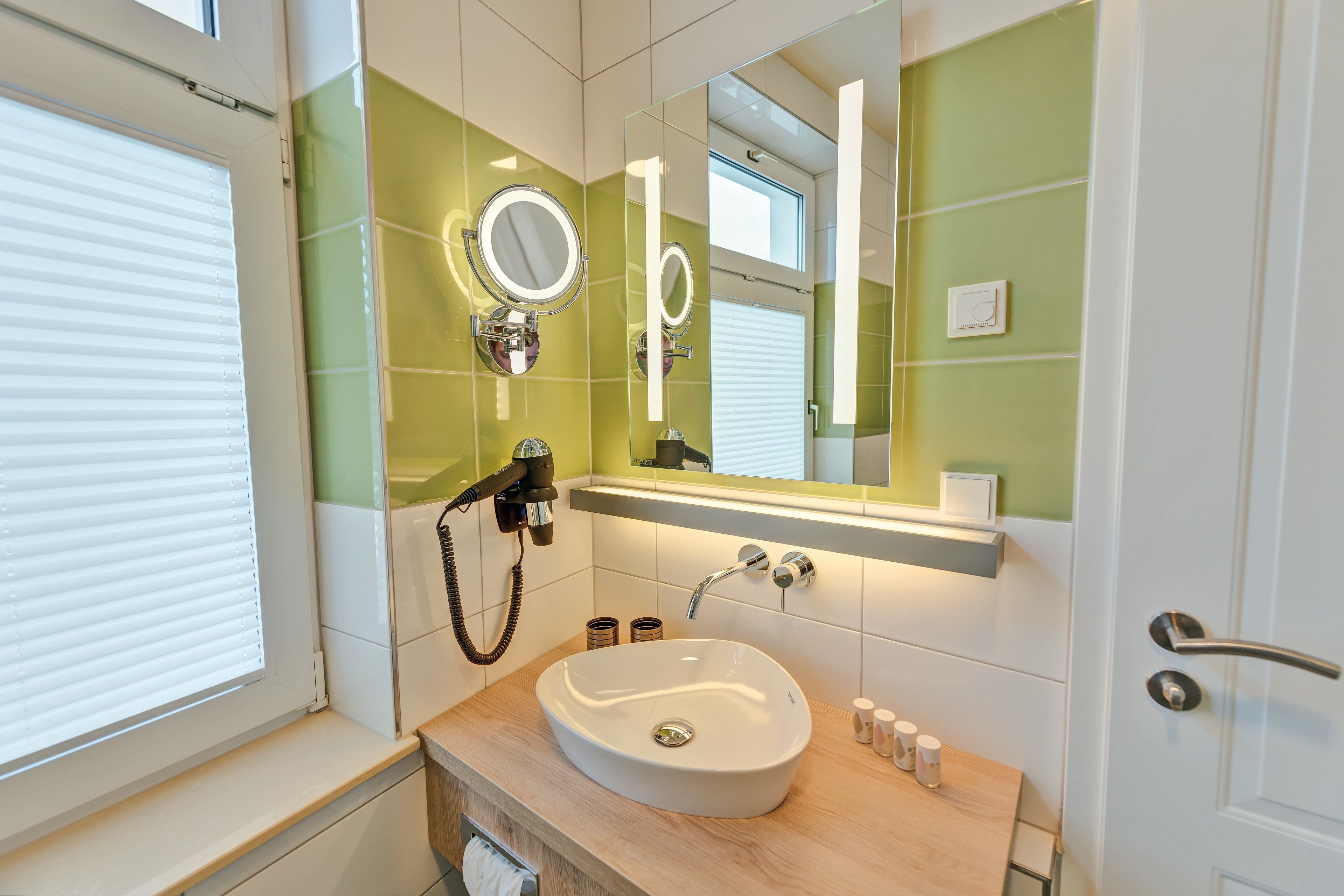 Baño con lavamanos blanco, paredes con azulejos verdes, espejo con luces, toallero y ventana con persianas.
