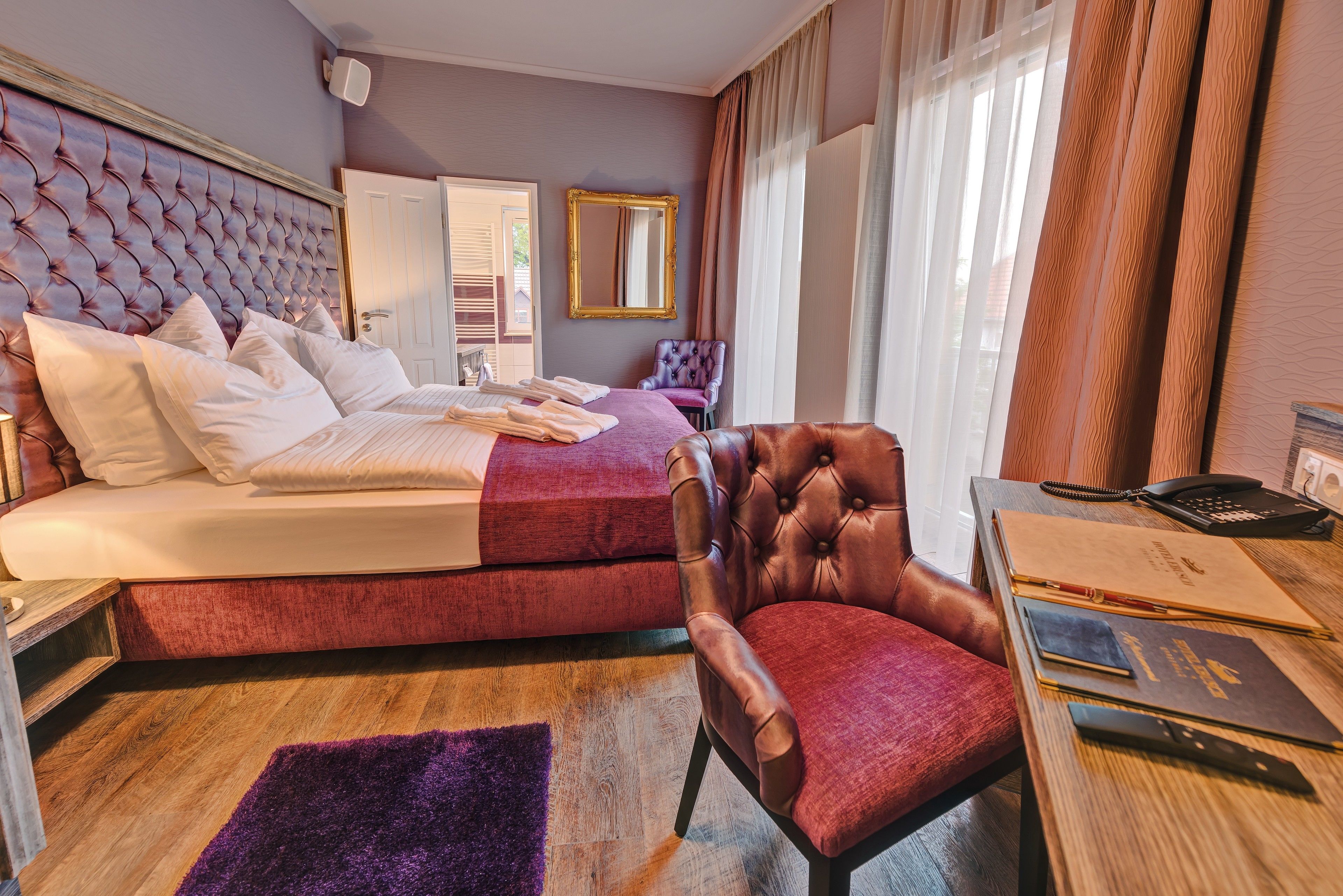 Una habitación de hotel con una cama grande, una silla, un escritorio con teléfono y documentos, y una ventana con cortinas.