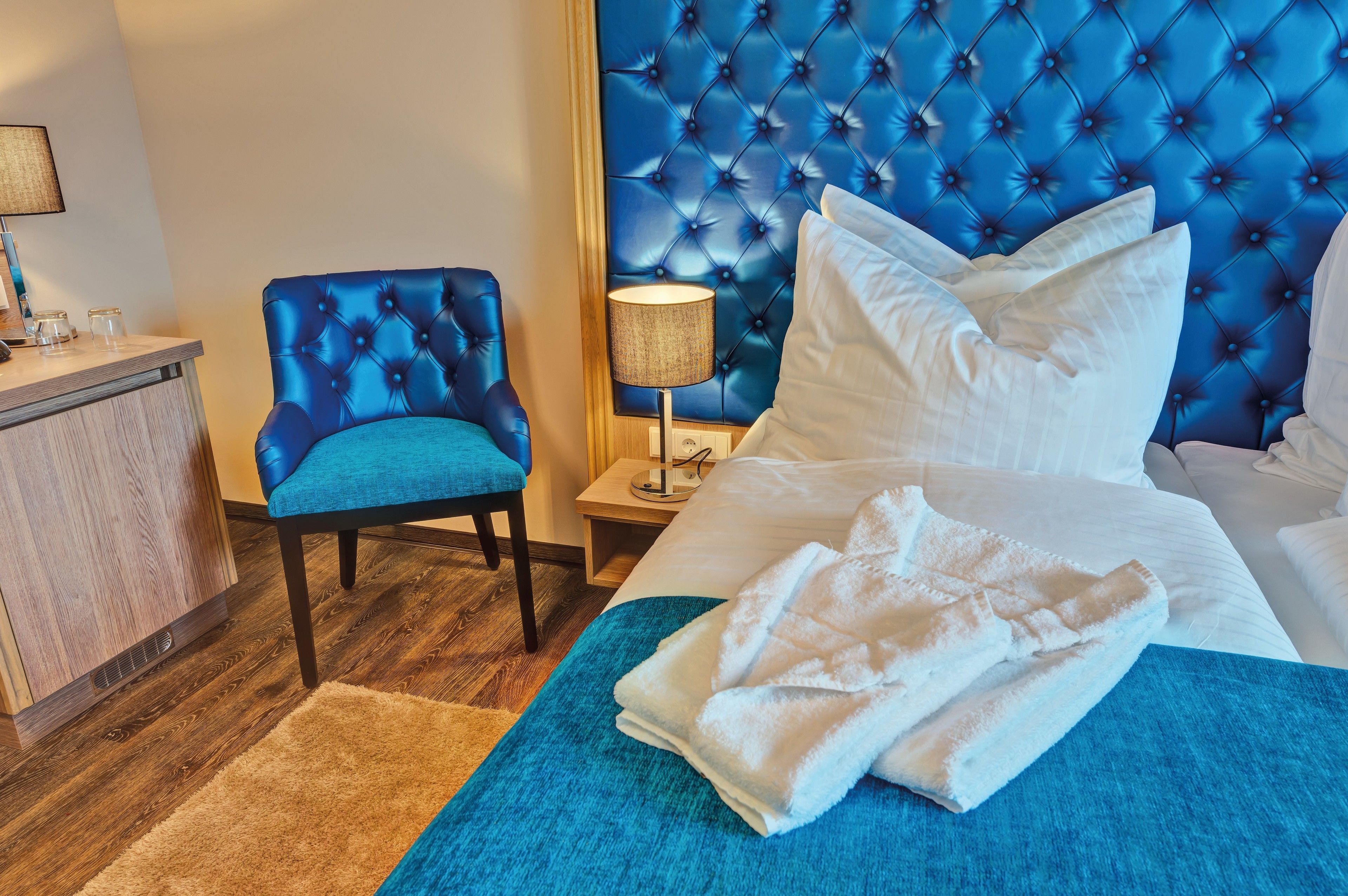 A hotel room with a tufted headboard, a blue upholstered chair, a bedside table with a lamp, and a neatly made bed with a white blanket.