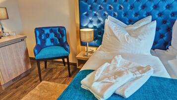 A hotel room with a tufted headboard, a blue upholstered chair, a bedside table with a lamp, and a neatly made bed with a white blanket.