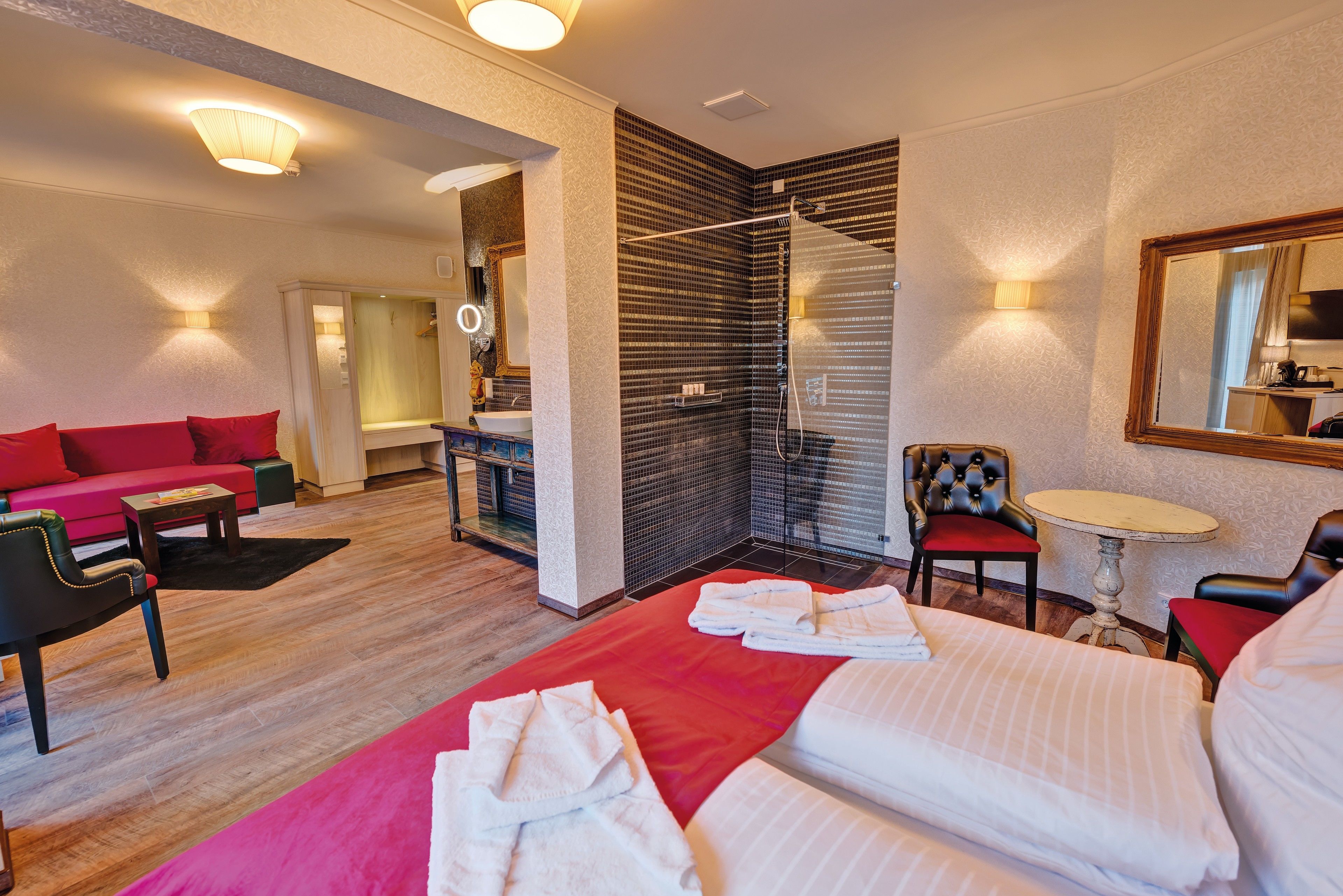 Et hotelværelse med seng, rød sofa, lille bord, stol og bruseområde.
