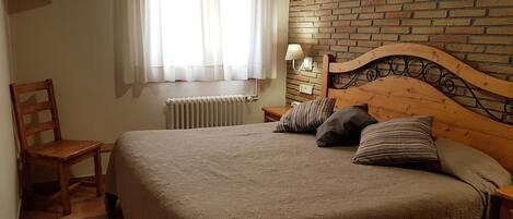 A bedroom with a wooden headboard, a bed with patterned bedding, a chair, a TV mounted on the wall, and a window with curtains.