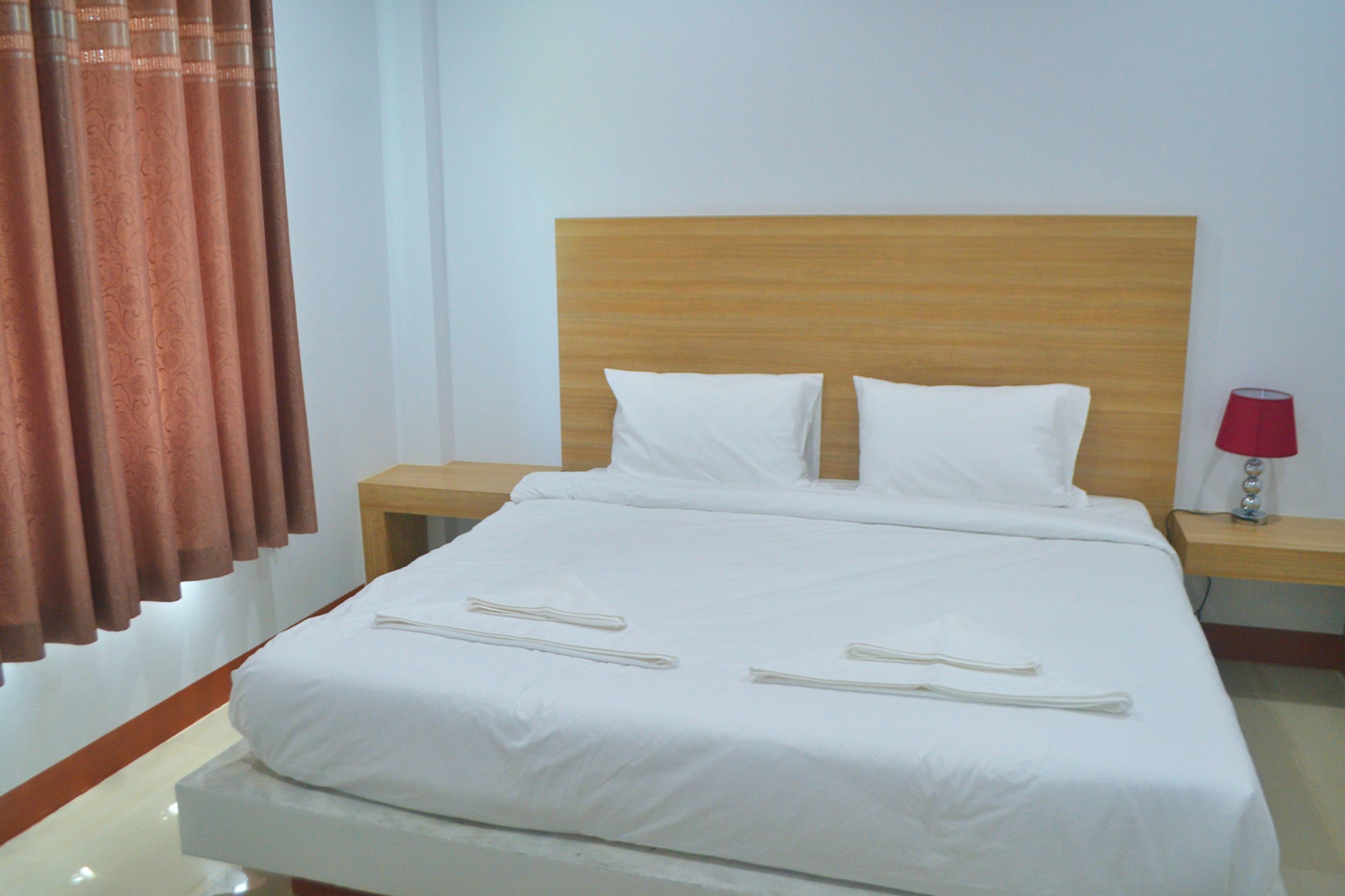 A neatly made bed with white linens, two pillows, and a wooden headboard. There are two bedside tables with lamps, one red and one white.