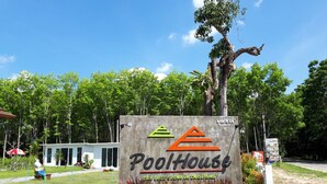 Segnale per una dependance della piscina con un logo, circondata da vegetazione e un edificio sullo sfondo.