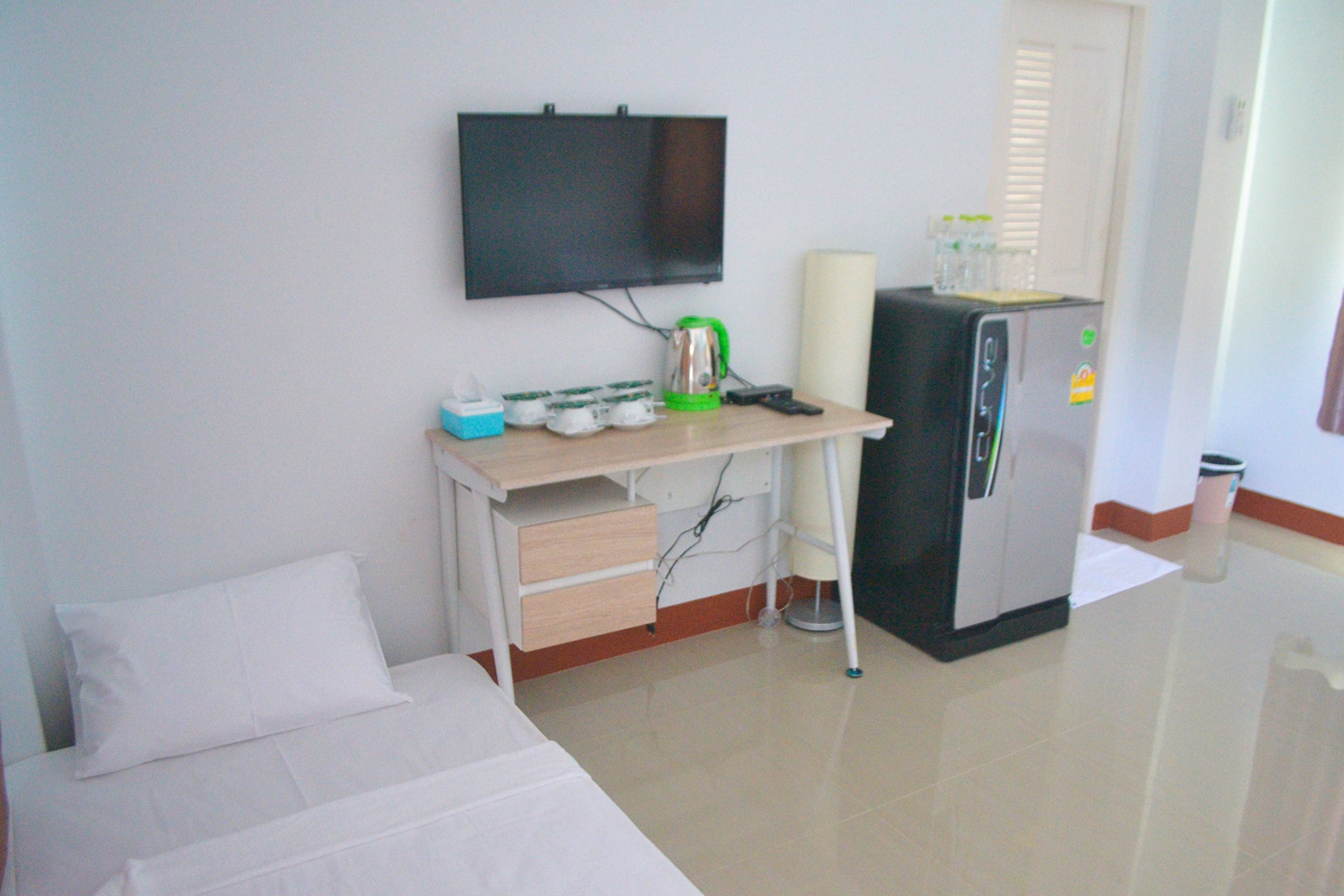 A hotel room with a bed, a desk with a television, a refrigerator, and a trash bin.