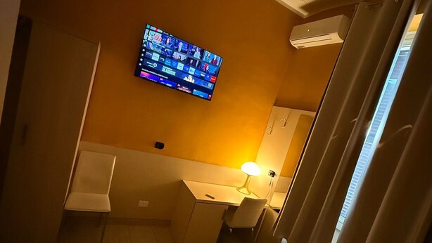 A hotel room with a bed, a desk, a chair, a TV mounted on the wall, and an air conditioning unit. - Hola Roma (Rome)