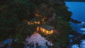 Une propriété isolée située en bord de mer, avec un bâtiment doté d’un toit en verre, entouré d’une forêt dense et offrant une vue sur une côte rocheuse.