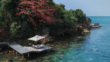 Una zona costera con un muelle de madera que se adentra en aguas azul turquesa, rodeada de exuberante vegetación y un acantilado rocoso.